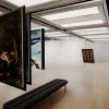 展示室に吊り下げられた複数の絵画と手前のベンチ